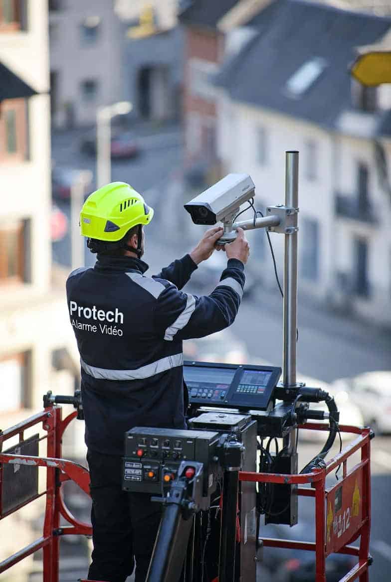 Technicien de Protech Alarme Vidéo en nacelle réglant une caméra de vidéosurveillance vue aérienne sur les toits d’une ville avec bâtiments anciens.