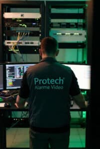 Homme de dos devant plusieurs écrans de supervision, portant un gilet sombre floqué « Protech Alarme Vidéo », travaillant dans une salle serveur avec baies informatiques éclairées en vert.