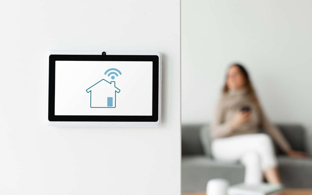 Une tablette murale blanche affichant l'icône d'une maison connectée avec des ondes Wi-Fi bleues au-dessus du toit, dans un salon moderne où une femme assise sur un canapé gris tient un smartphone.